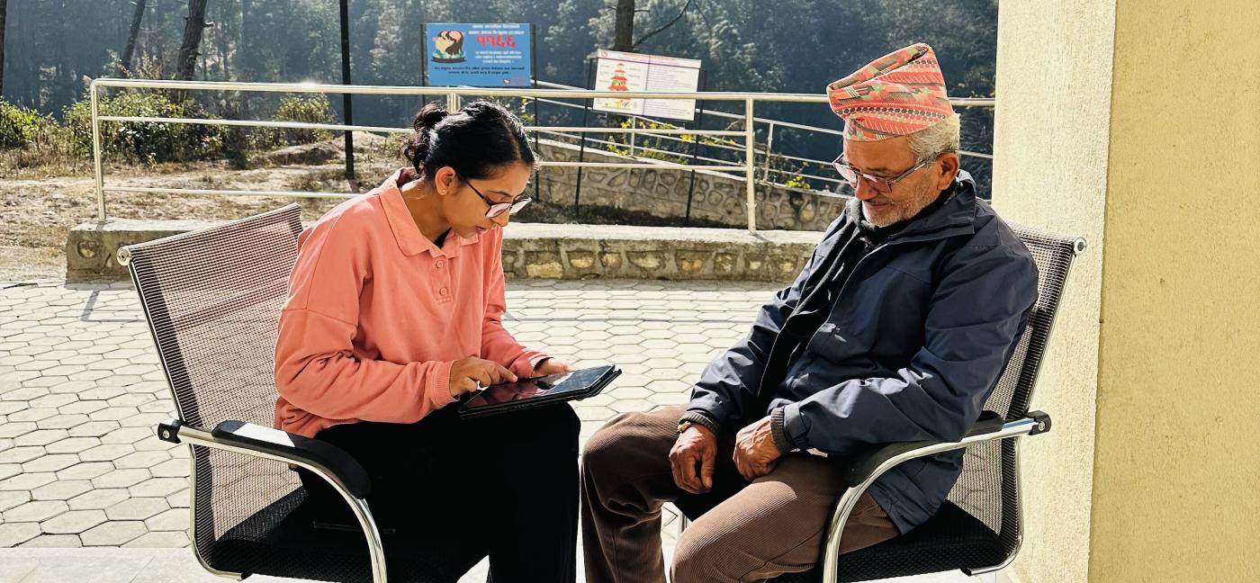 A young woman in a pink shirt interacts with an elderly man in a traditional Nepali cap and jacket as they sit on modern chairs outdoors. Behind them, a serene forest landscape and a bright blue sky create a tranquil atmosphere