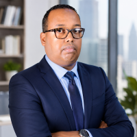 Professional headshot of Djibril Moussa wearing a navy suit, light blue shirt, and navy tie, with arms crossed against a blurred office background.