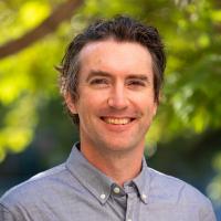 Portrait of Niall Kavanagh smiling at the camera outdoors, wearing a light blue button-down shirt. A softly blurred background of green trees and warm sunlight creates a bright, natural setting.