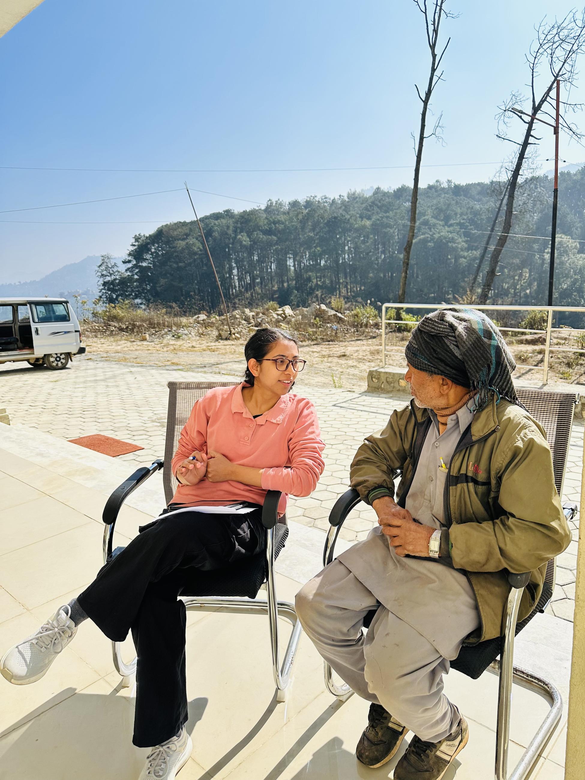 A young woman in a pink shirt and glasses sits cross-legged in a chair, holding a pen and paper as she engages in conversation with an elderly man wearing a green jacket and a traditional headwrap. They are seated outdoors on a sunny day, with a forested hillside and a parked van visible in the background