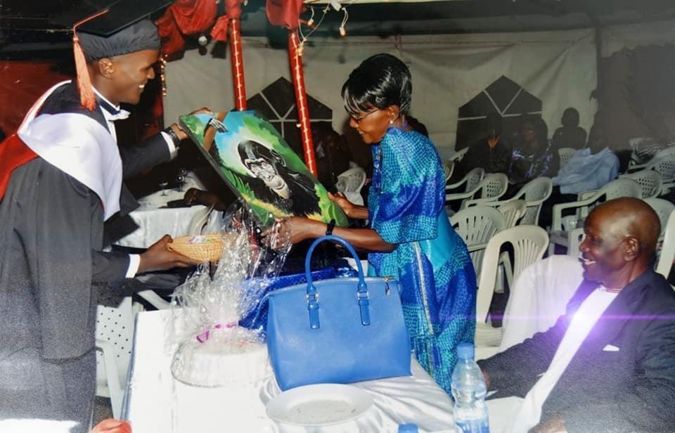 A young man in graduation attire presents a colorful portrait to an older woman at a celebration, while others look on and smile.