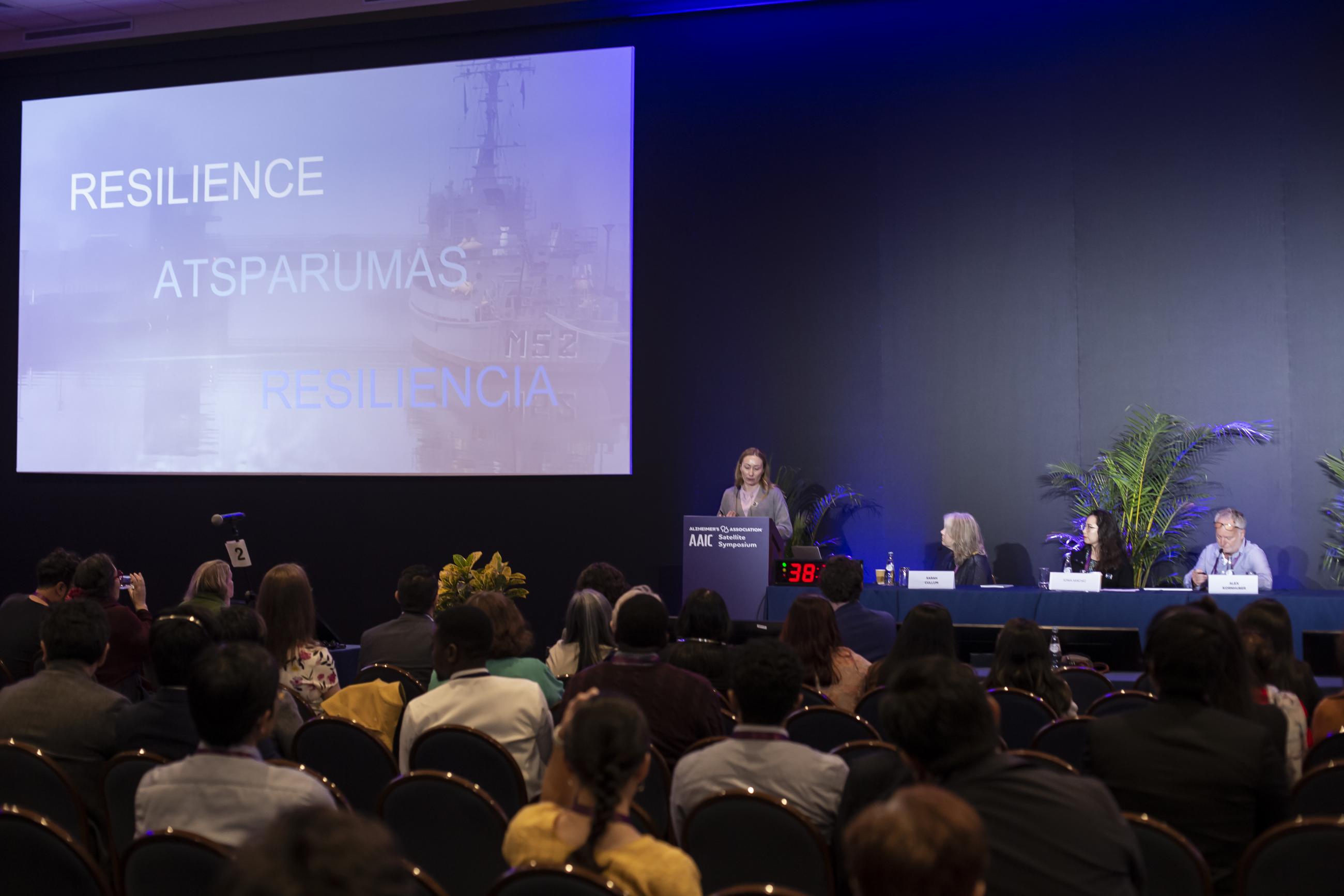 A woman speaks at a podium during a conference, with the word “Resilience” displayed behind her in multiple languages.