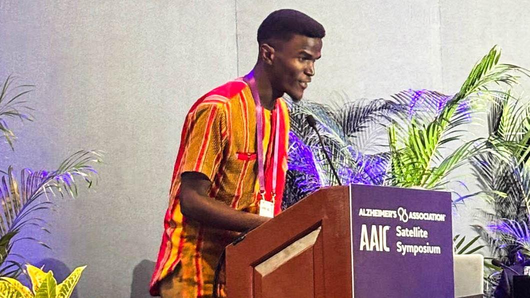 A man speaks at a podium during the AAIC Satellite Symposium, with a visible countdown timer and plants in the background.
