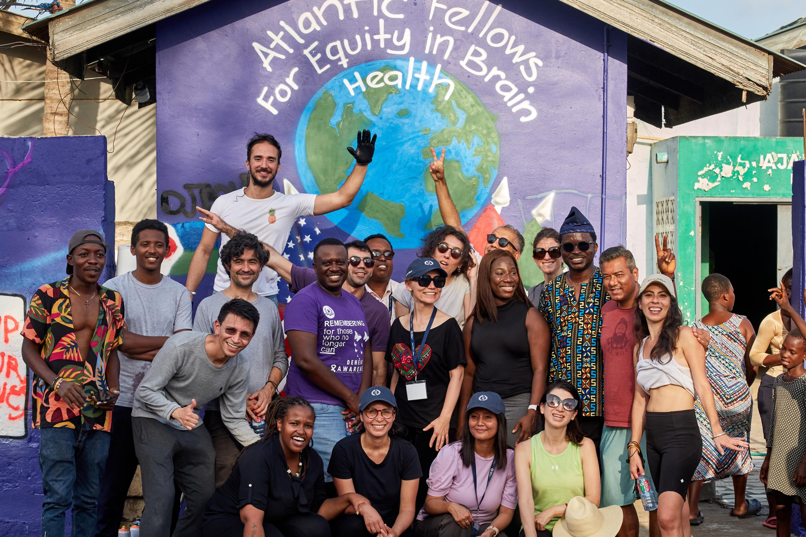 A diverse group of Atlantic Fellows smiling and posing together in front of a colorful mural with the words ‘Atlantic Fellows for Equity in Brain Health’ painted above a globe.