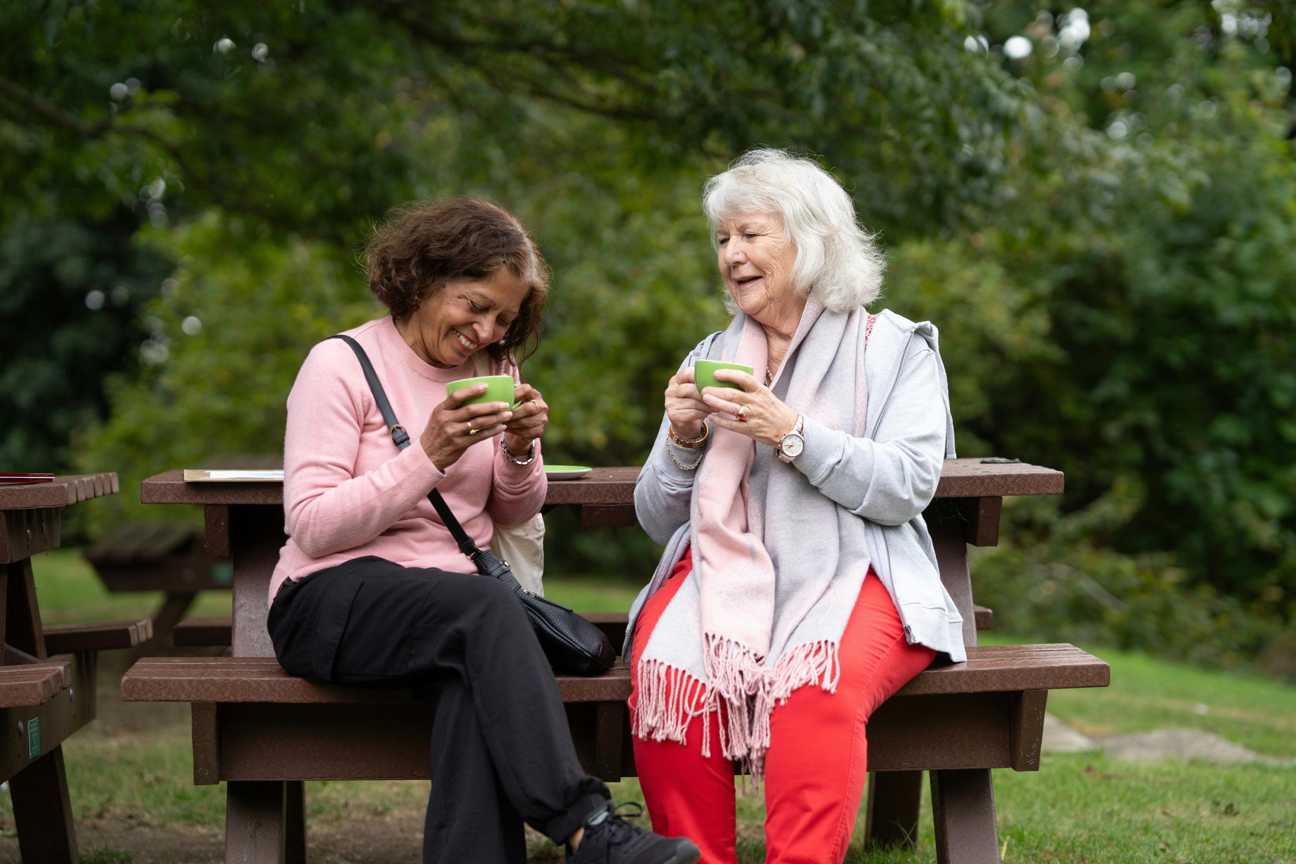 Two older women sitting on a bench outdoors, smiling and enjoying hot drinks together.”