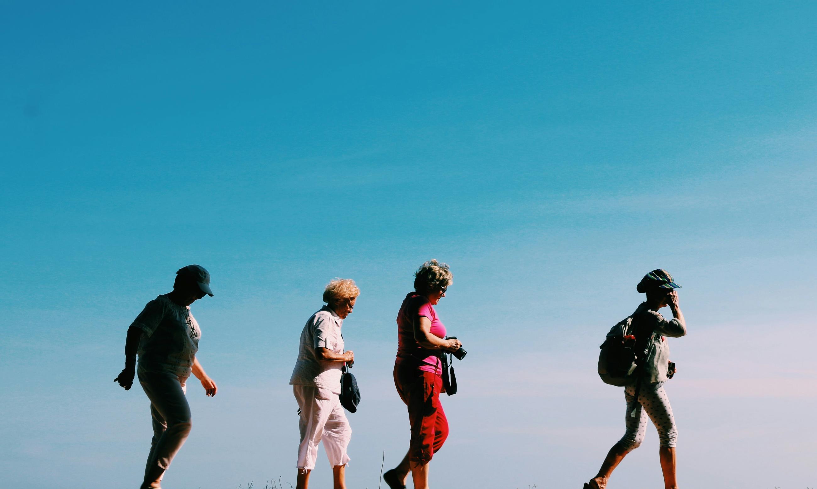 Four people are walking in a line outdoors on a sunny day. They are wearing hats and casual clothing, some carrying small bags or a backpack, with a clear blue sky in the background.