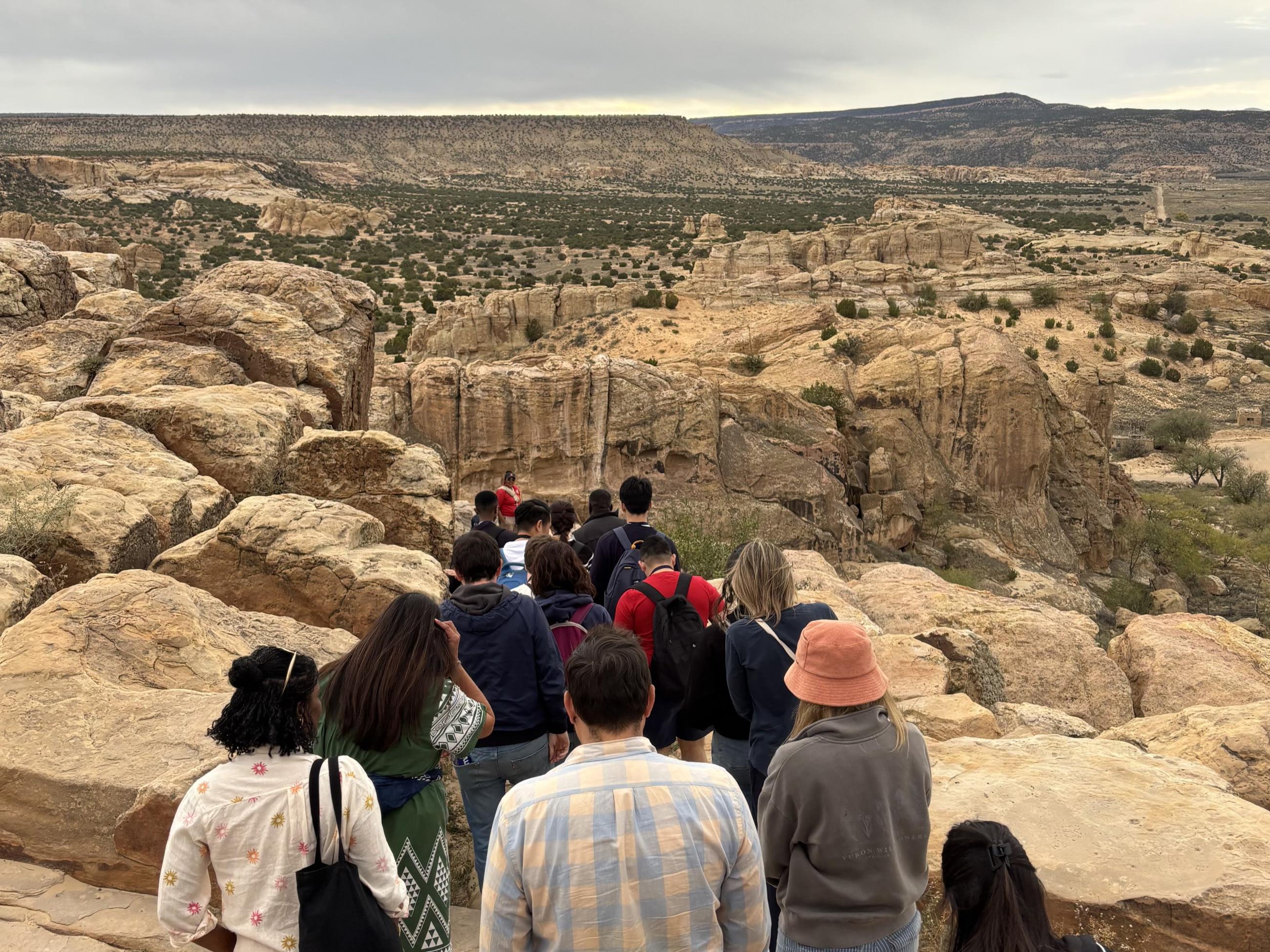 A group of Atlantic Fellows walk along a rocky trail overlooking the New Mexico landscape.