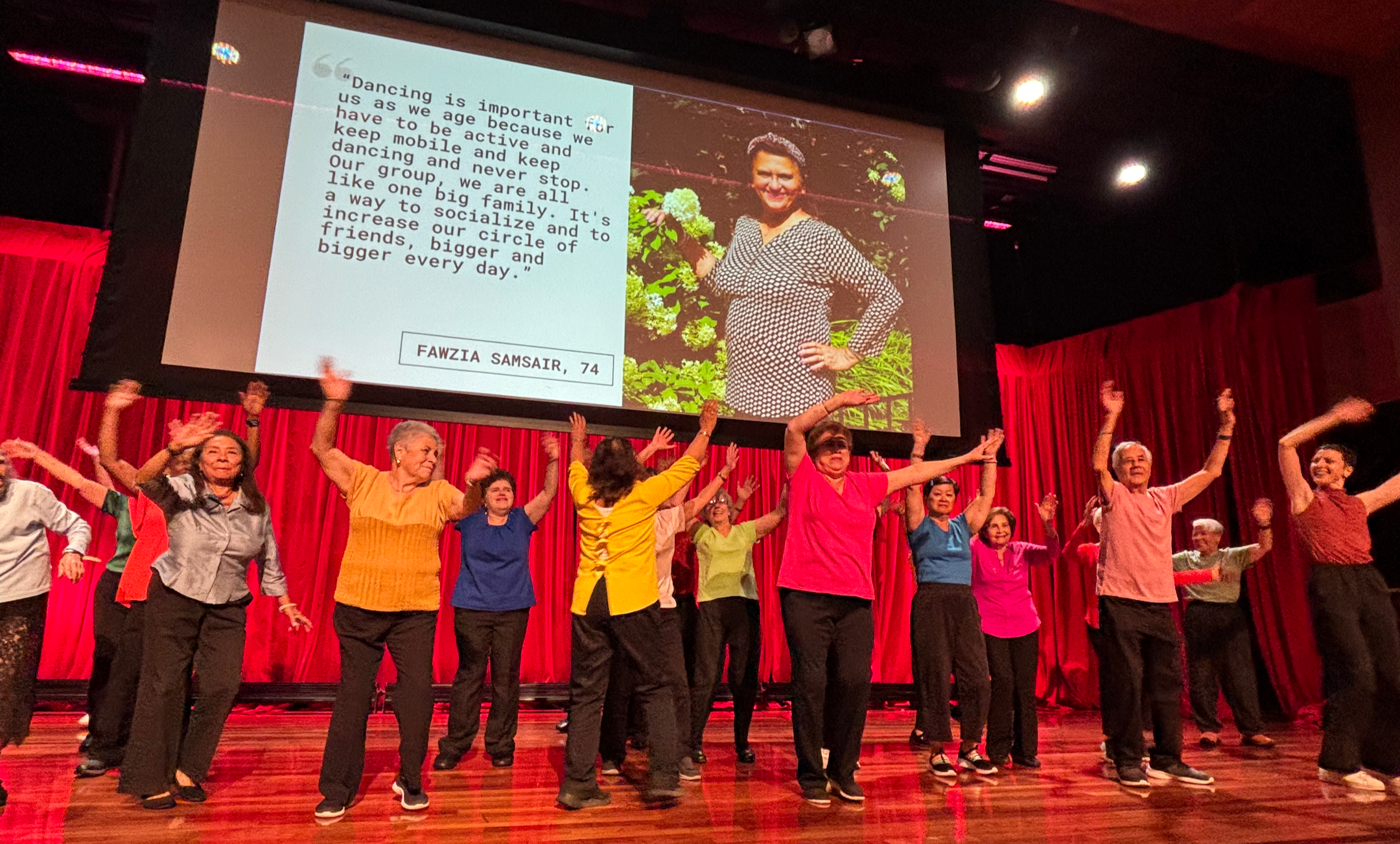 A group of older adults dance together on stage with arms raised in celebration.