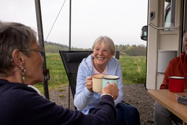 Group of senior friends on a hiking staycation, staying in a camper van in Dumfries and Galloway, Scotland. They are sitting outside enjoying lunch, drinking tea, toasting their mugs.