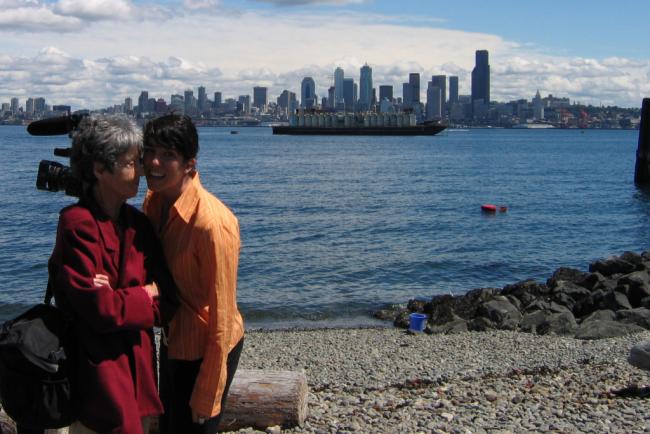 Two people stand close together and smile near a rocky shoreline, with a city skyline and a large cargo ship visible across the water in the background.