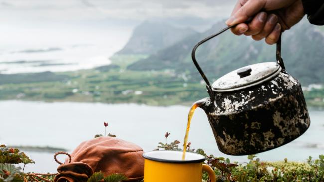A person pours tea from a rustic kettle into a yellow mug while sitting on a grassy hillside with mountains and water in the background.