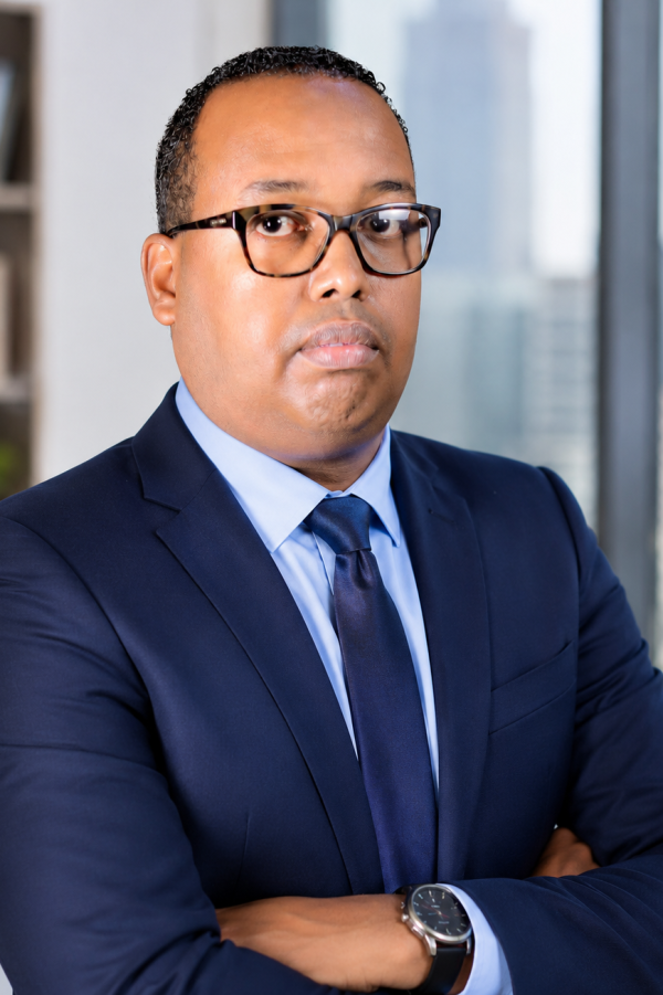 Professional headshot of Djibril Moussa wearing a navy suit, light blue shirt, and navy tie, with arms crossed against a blurred office background.