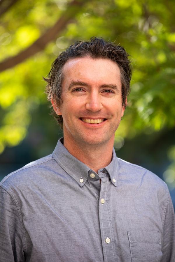 Portrait of Niall Kavanagh smiling at the camera outdoors, wearing a light blue button-down shirt. A softly blurred background of green trees and warm sunlight creates a bright, natural setting.