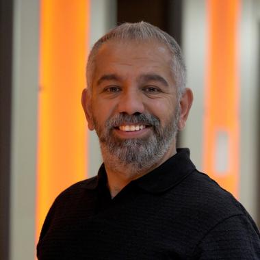 Armağan Uysal smiles at the camera, wearing a black polo shirt and light trousers.