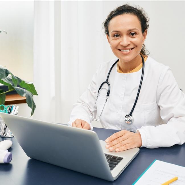 Smiling general practitioner sits at her workplace wearing a medical coat with a laptop.
