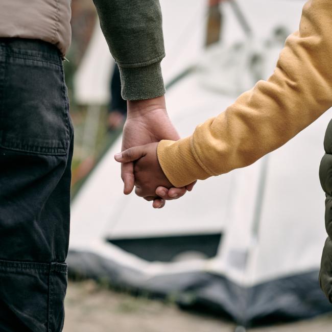 Rear view of unrecognizable parent of elder brother holding hand of child against tent in migrant camp