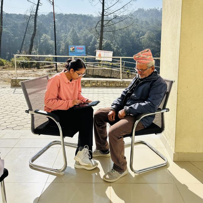 A young woman in a pink shirt interacts with an elderly man in a traditional Nepali cap and jacket as they sit on modern chairs outdoors. Behind them, a serene forest landscape and a bright blue sky create a tranquil atmosphere