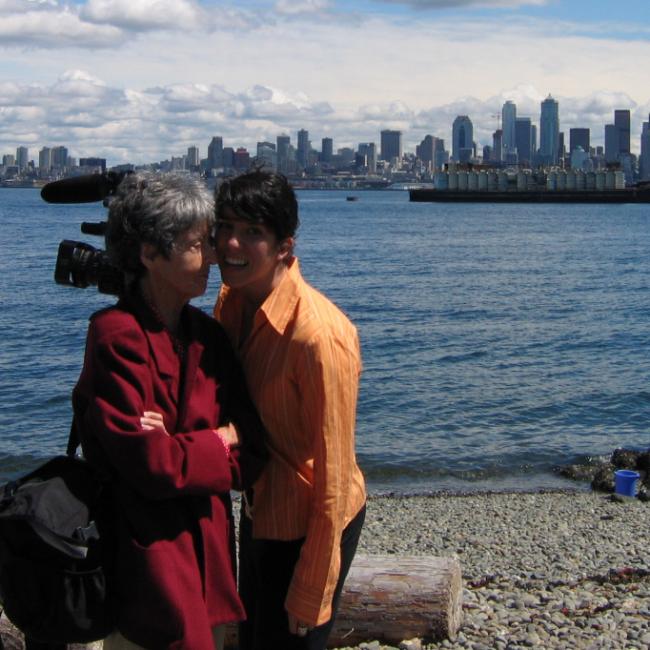Two people stand close together and smile near a rocky shoreline, with a city skyline and a large cargo ship visible across the water in the background.