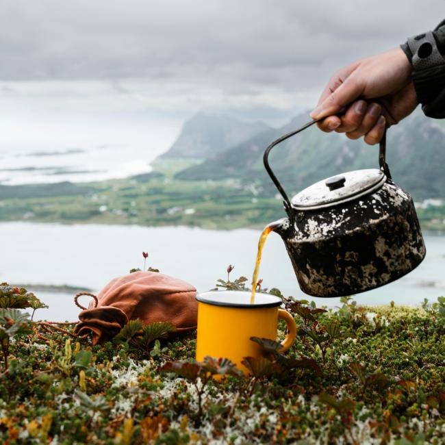A person pours tea from a rustic kettle into a yellow mug while sitting on a grassy hillside with mountains and water in the background.