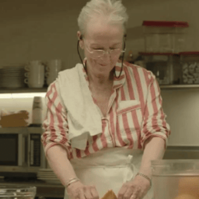 A scene from the film &quot;Familiar Touch&quot; shows an older woman in a striped shirt, apron, and headset preparing food in a kitchen.