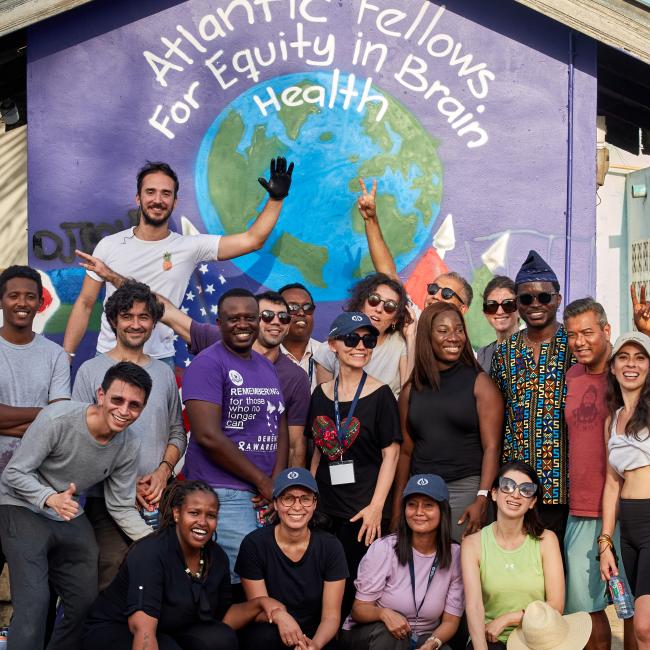 A diverse group of Atlantic Fellows smiling and posing together in front of a colorful mural with the words ‘Atlantic Fellows for Equity in Brain Health’ painted above a globe.