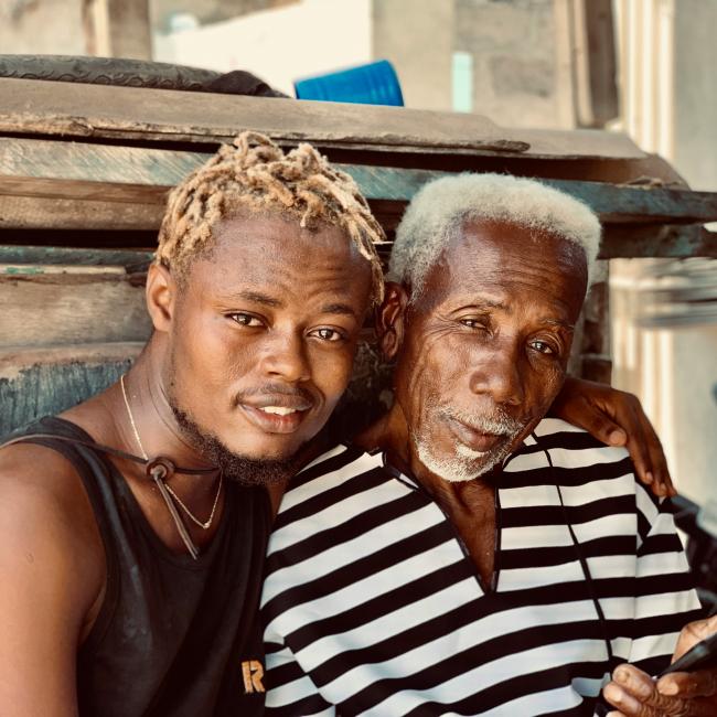 A young man with light blond twists sits closely beside an older man with white hair. They are smiling gently and leaning toward each other, showing warmth and connection.