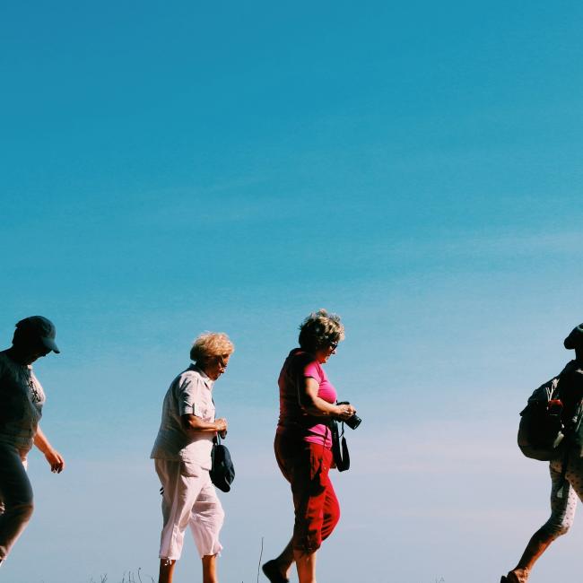 Four people are walking in a line outdoors on a sunny day. They are wearing hats and casual clothing, some carrying small bags or a backpack, with a clear blue sky in the background.
