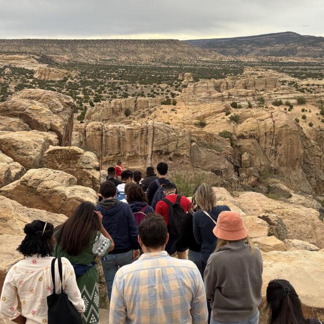 A group of Atlantic Fellows walk along a rocky trail overlooking the New Mexico landscape.