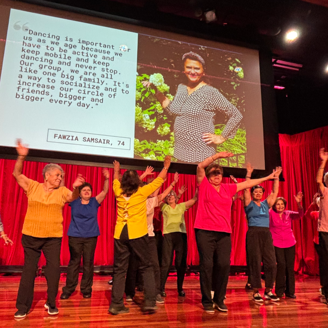 A group of older adults dance together on stage with arms raised in celebration.