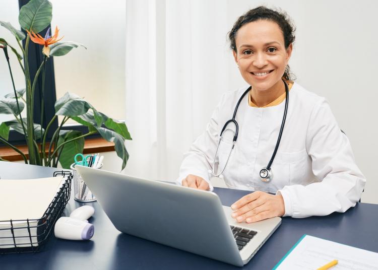 Smiling general practitioner sits at her workplace wearing a medical coat with a laptop.