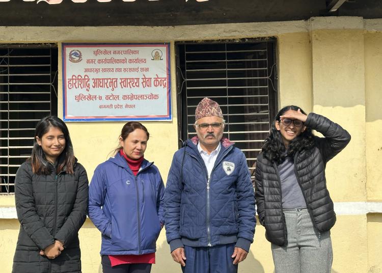 Four individuals pose in front of the Harisiddhi Ayurvedic Health Center in Dhulikhel, Nepal. The background features a yellow building with a red tiled roof and a signboard in Nepali script. Two women wear jackets, a man in a traditional Nepali hat stands center, and a young woman shields her eyes from the sun with a playful gesture