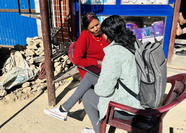 Two women engage in a heartfelt conversation outside a blue-doored local shop. One wears a vibrant red sweater, leaning slightly forward as she listens intently. The other, seated with a gray backpack, holds a tablet and speaks thoughtfully. A spiral staircase, brick wall, and scattered stones frame the sunlit scene