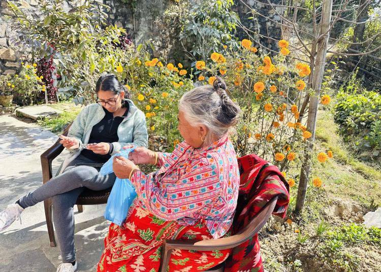 Two women sit outside surrounded by vibrant yellow marigolds and lush greenery. One woman, wearing a colorful patterned outfit, hands over a small blue bag while the other woman examines its contents with a smile. The bright sunlight and floral backdrop create a warm and lively atmosphere