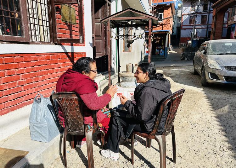 Two women sit on plastic chairs in a narrow alleyway, engaged in a lively conversation. One woman, wrapped in a maroon shawl, gestures animatedly while the other listens attentively, smiling. Behind them, a red-brick wall and a small shrine add character to the scene, with a silver car and colorful houses visible in the background