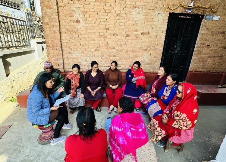 A young woman in a blue jacket sits on a colorful stool, passionately leading a discussion with a group of women seated in a semicircle. The women, dressed in vibrant traditional clothing, are engaged in the conversation. The setting features a rustic brick wall adorned with a garland and a black wooden door, evoking a sense of cultural connection and collaboration