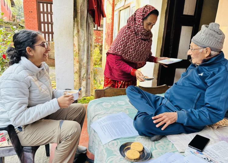 A woman in a white jacket smiles while holding a cup of tea during a conversation with an elderly man wearing a gray beanie and blue tracksuit, seated on a table. Another woman in a red patterned shawl serves tea on a tray. Papers, cookies, and a smartphone are placed on the table in a bright, cozy courtyard setting