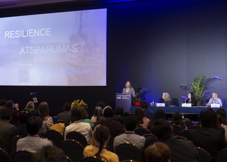 A woman speaks at a podium during a conference, with the word “Resilience” displayed behind her in multiple languages.
