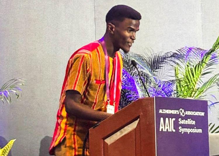 A man speaks at a podium during the AAIC Satellite Symposium, with a visible countdown timer and plants in the background.