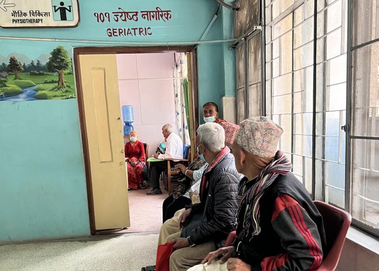 Elderly people sit on chairs in a hospital waiting area marked “Geriatric.”