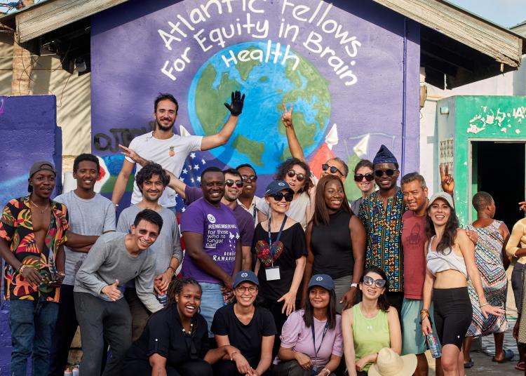 A diverse group of Atlantic Fellows smiling and posing together in front of a colorful mural with the words ‘Atlantic Fellows for Equity in Brain Health’ painted above a globe.