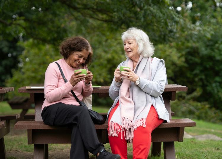 Two older women sitting on a bench outdoors, smiling and enjoying hot drinks together.”