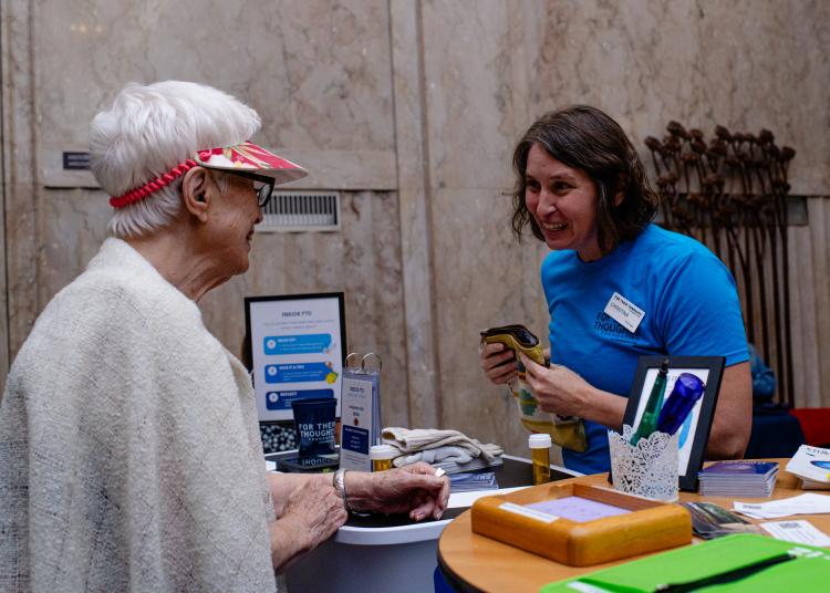 A woman in a blue shirt smiles while speaking with a visitor at an FTD information table during the UnRavelled event.