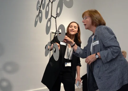 two women interact with a piece of hanging artwork