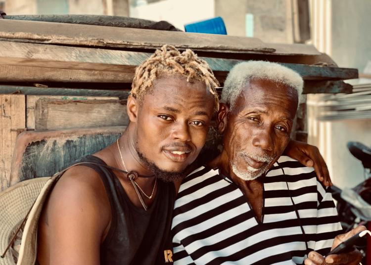 A young man with light blond twists sits closely beside an older man with white hair. They are smiling gently and leaning toward each other, showing warmth and connection.