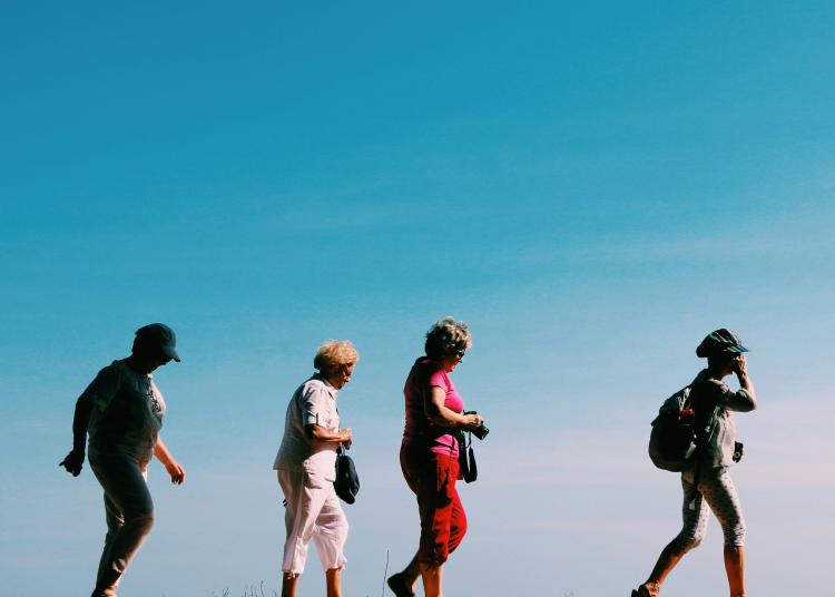 Four people are walking in a line outdoors on a sunny day. They are wearing hats and casual clothing, some carrying small bags or a backpack, with a clear blue sky in the background.