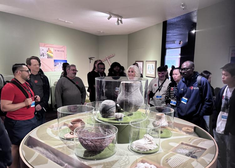Atlantic Fellows stand around a museum exhibit, listening and observing during a guided visit.