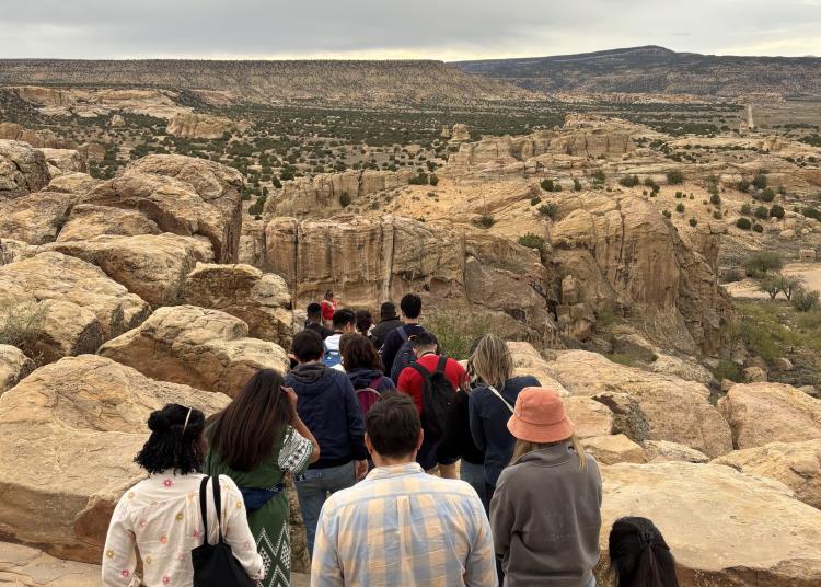 A group of Atlantic Fellows walk along a rocky trail overlooking the New Mexico landscape.