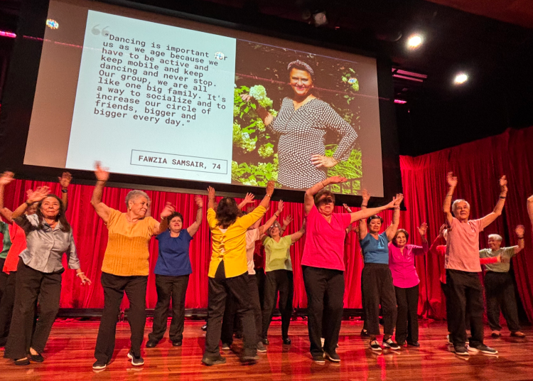 A group of older adults dance together on stage with arms raised in celebration.