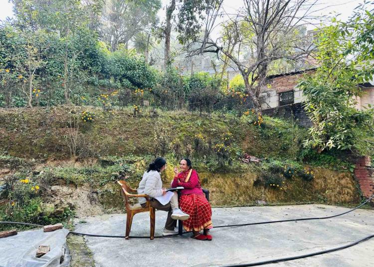 A healthcare worker sits with an older woman outdoors in Nepal, speaking and taking notes during a home visit as part of a community health assessment.