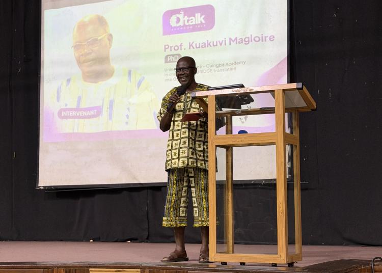 Magloire Kuakuvi speaks on stage in front of a projected Ahonhon Talk presentation about his translation work.